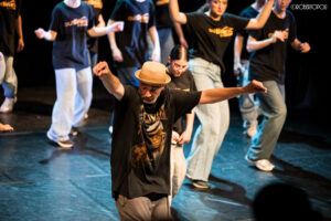 Gruppo di ballerini durante una coreografia Urban Hip Hop sul palco, con un danzatore in cappello e braccia aperte in primo piano.